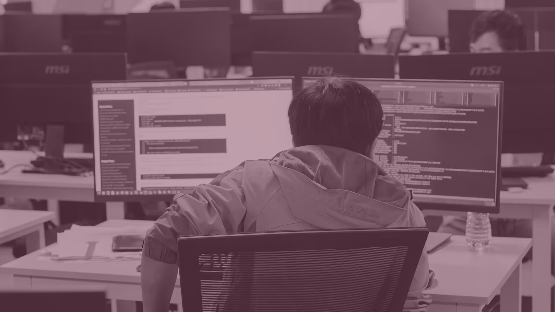 Person wearing a jacket working on two computer monitors displaying code and documentation in an office setting.