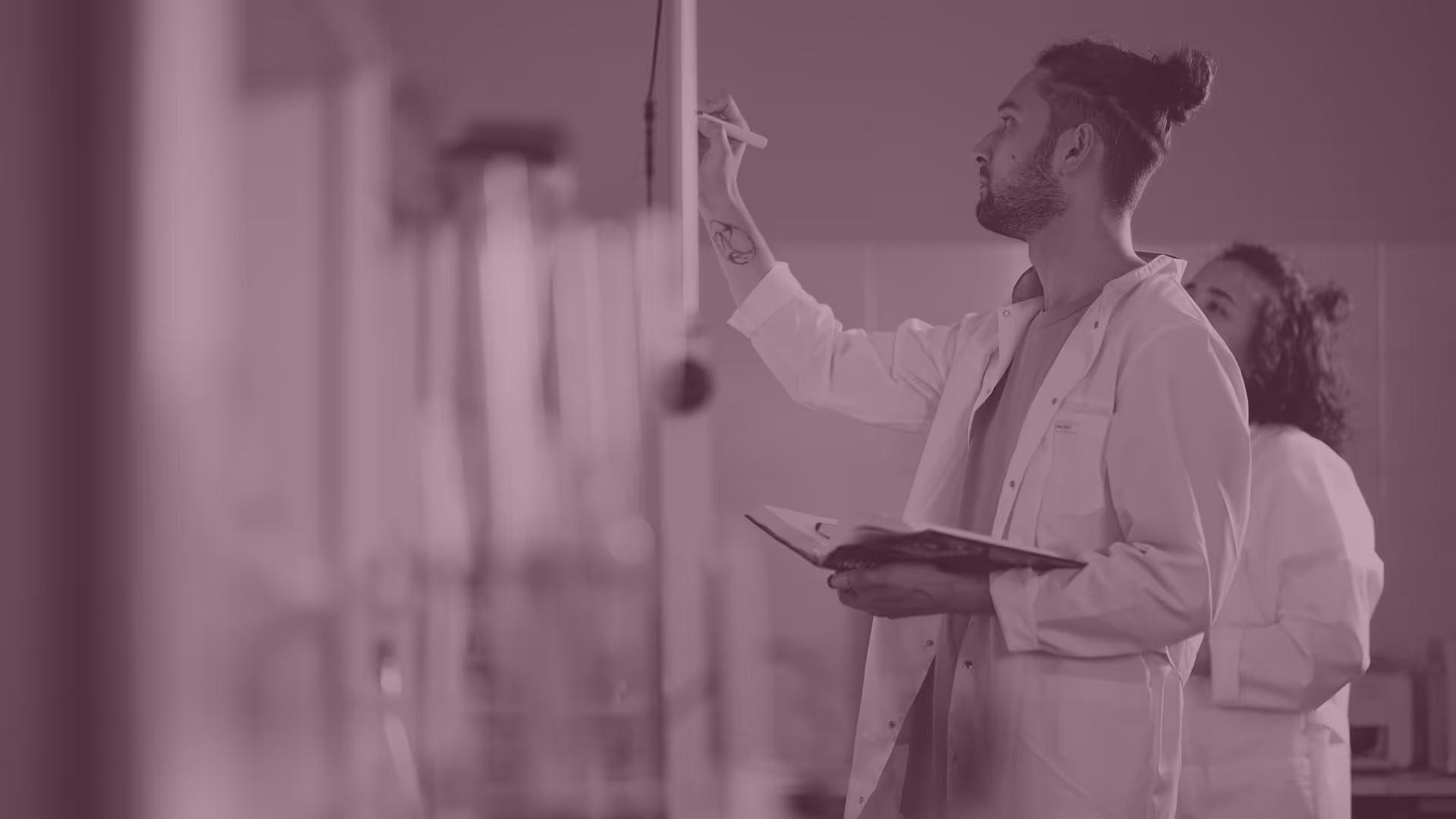 Scientist in a lab coat writing on a whiteboard while holding a book, with a colleague in lab coat in the background.