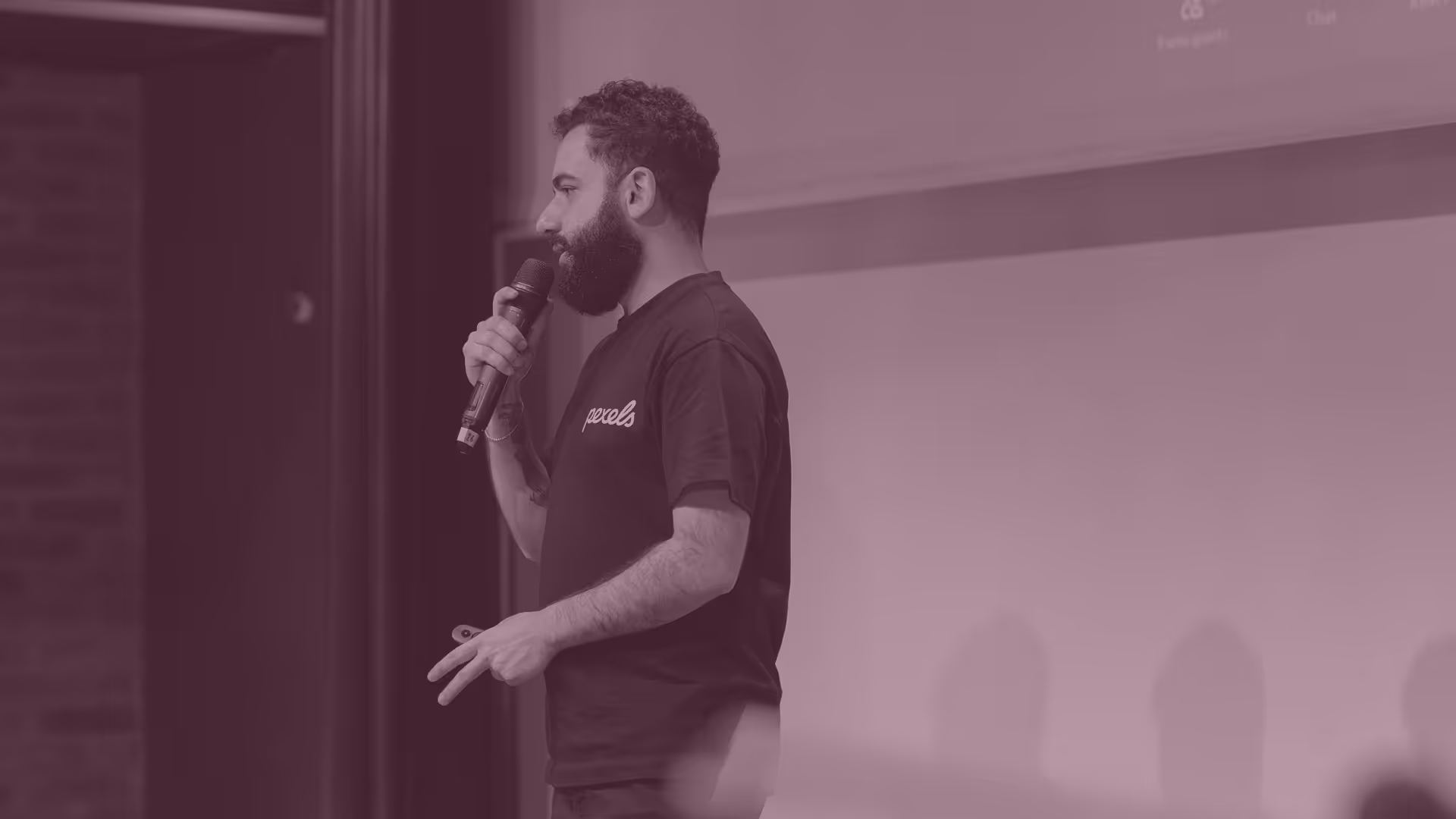 Bearded man in a black T-shirt holding a microphone and speaking on stage.