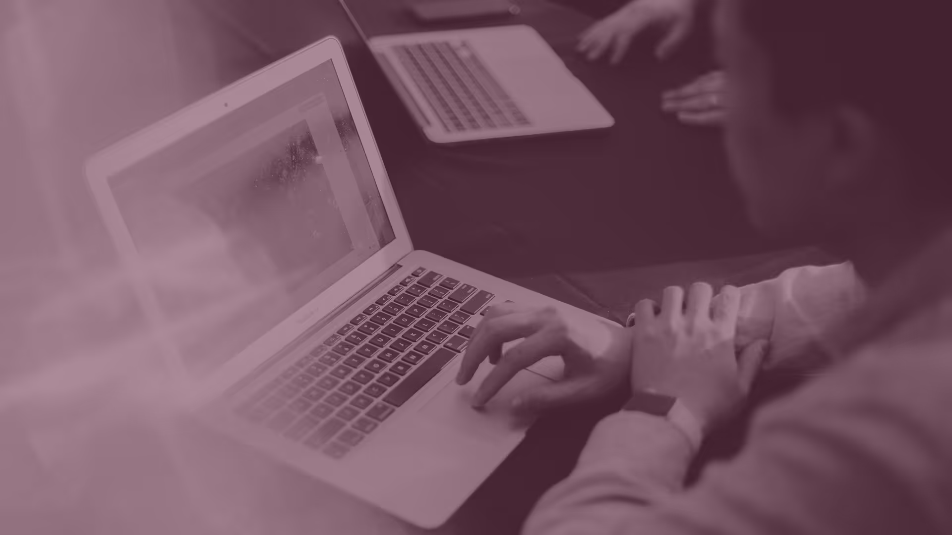 Person typing on a laptop keyboard with another laptop and someone else's hands visible on a table.