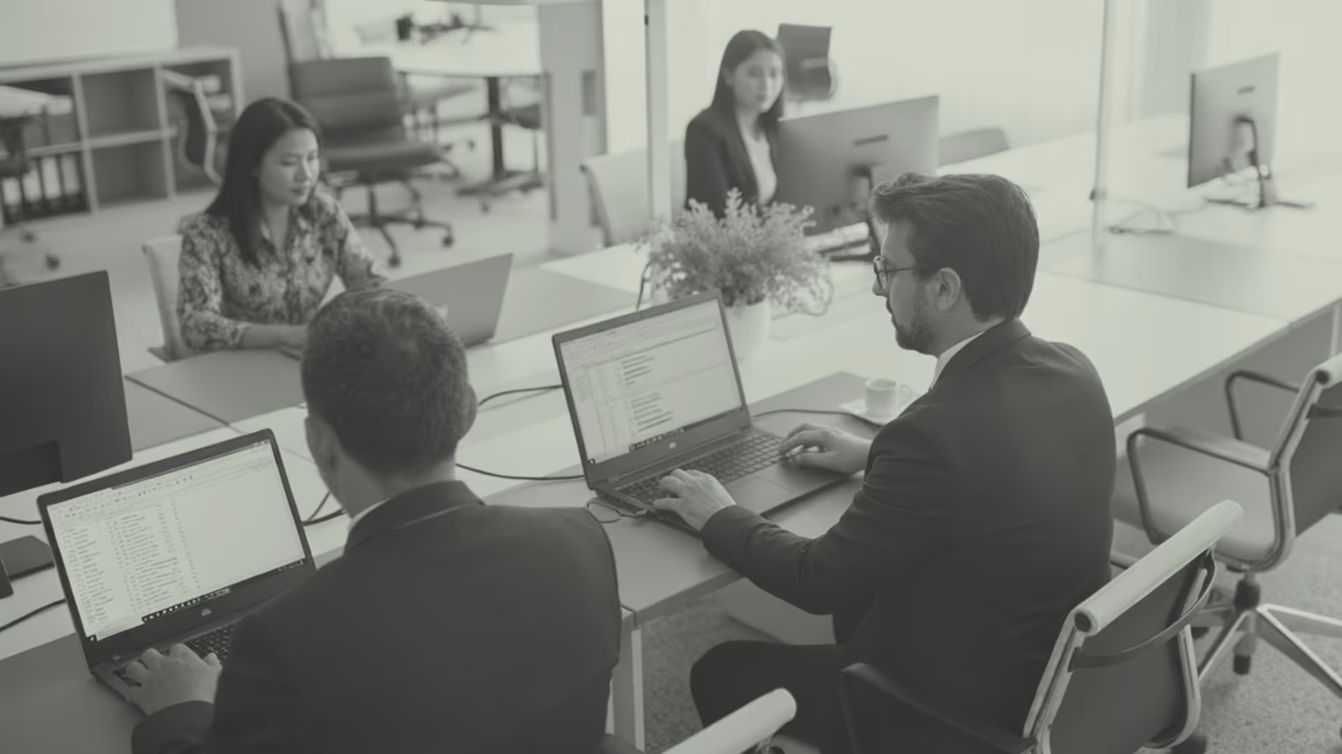 Four professionals working on laptops at a modern office desk with computers and office chairs.